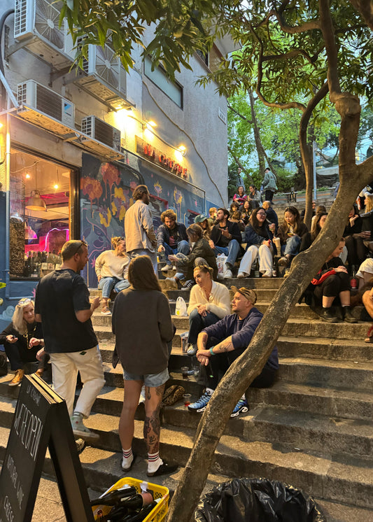People sitting on outdoor steps in an urban setting with a store and trees in the background.